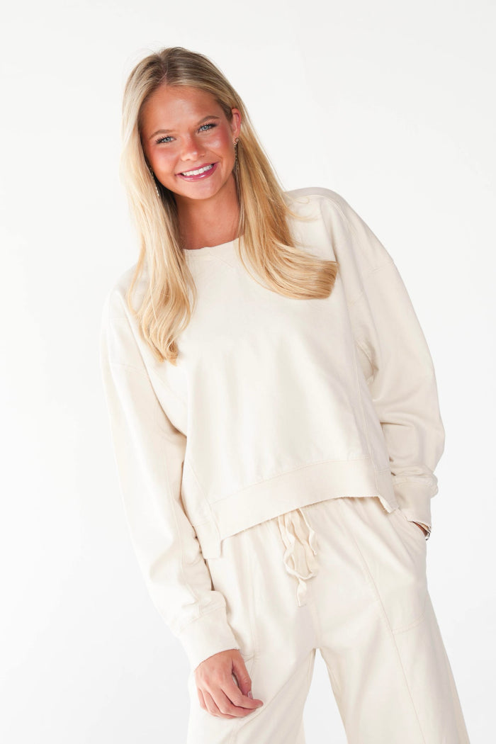 A smiling blonde woman wearing a matching cream-colored lounge set, featuring a cropped long-sleeve sweatshirt and drawstring sweatpants, set against a plain white studio background.