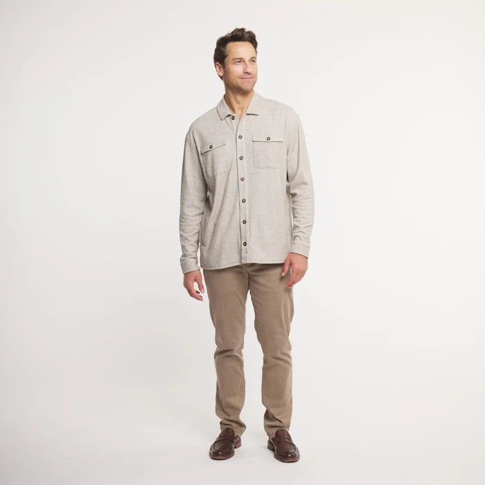 A man wearing a light beige long-sleeve button-up shirt with two chest pockets and tan pants, standing against a plain white studio background.
