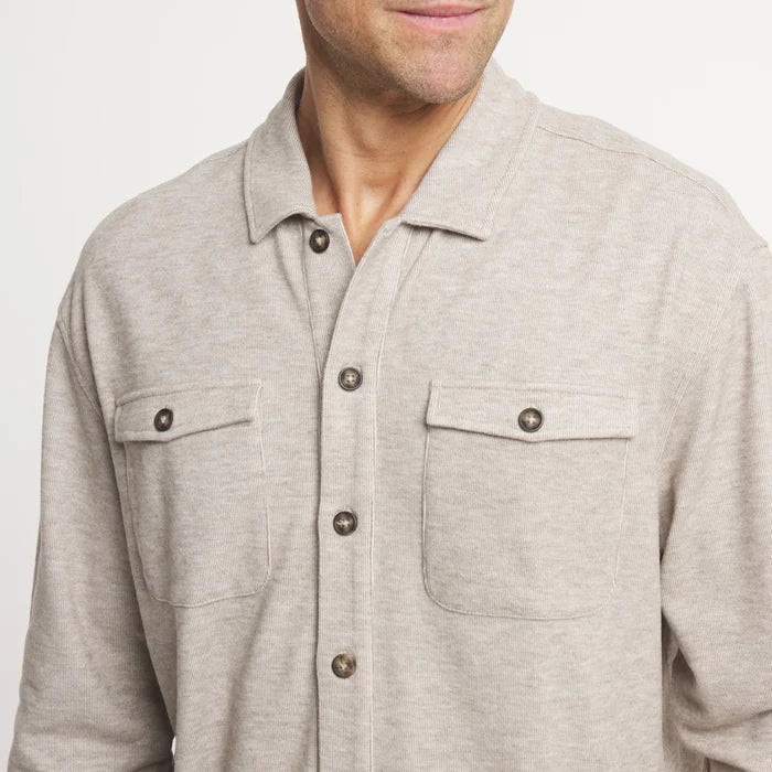 A close-up of a man wearing a light heather gray long-sleeve button-up shirt with two chest pockets and dark buttons, against a plain light background.