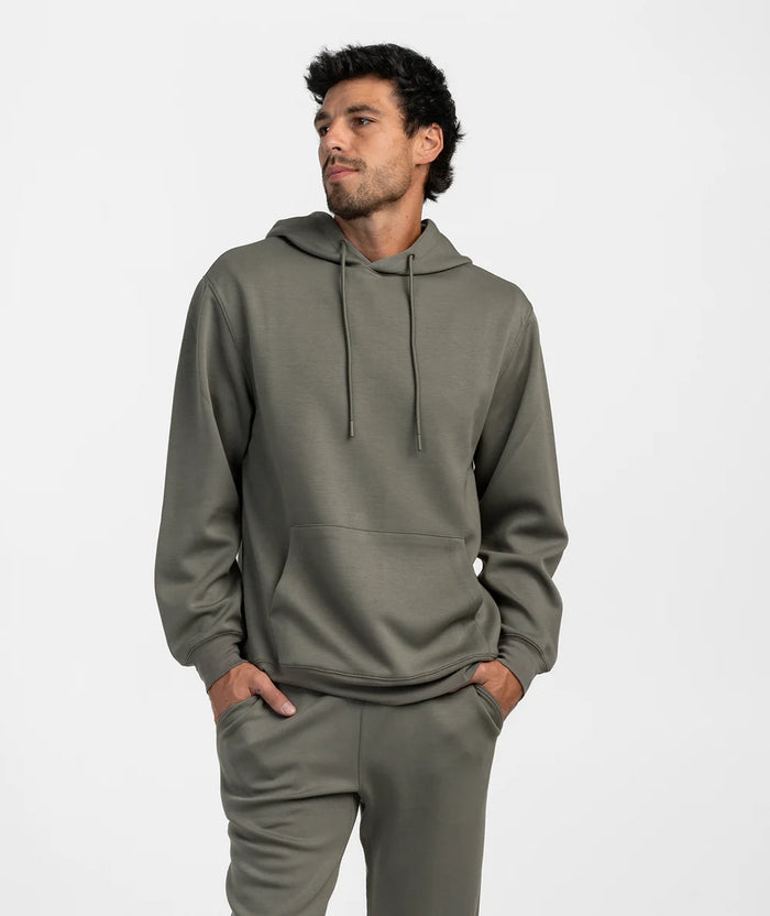 A man wearing a matching two-piece lounge set in a muted olive green color, featuring a pullover hoodie with drawstrings and matching sweatpants, set against a plain light gray background.