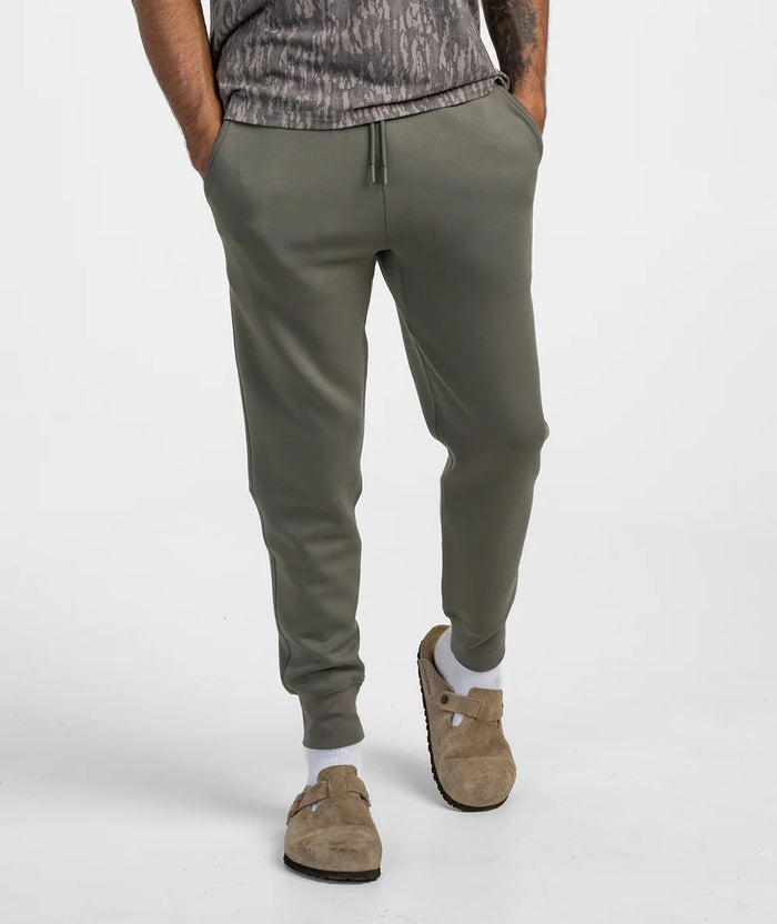 A person wearing olive green jogger sweatpants with a drawstring waist, paired with a gray patterned t-shirt, white socks, and brown clog-style shoes, shown from the waist down against a light gray studio background.