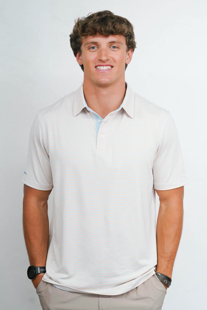 A smiling man wearing a light beige short-sleeve polo shirt with a subtle textured pattern, paired with khaki pants, standing against a plain light gray background.