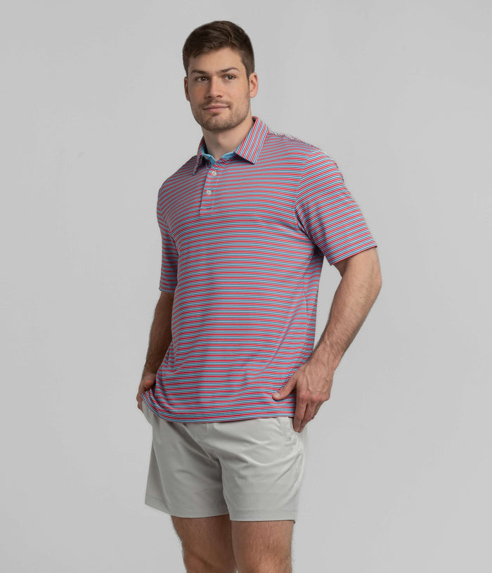 A man wearing a short-sleeve polo shirt with thin horizontal red, blue, and white stripes, paired with light gray shorts, standing against a solid gray studio background.