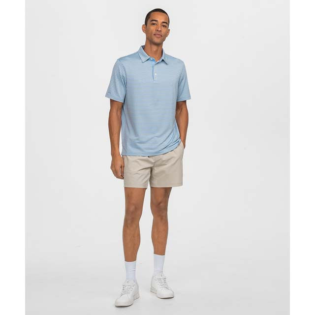 A man modeling a light blue short-sleeve polo shirt with beige shorts, white socks, and white sneakers, standing against a plain light gray studio background.