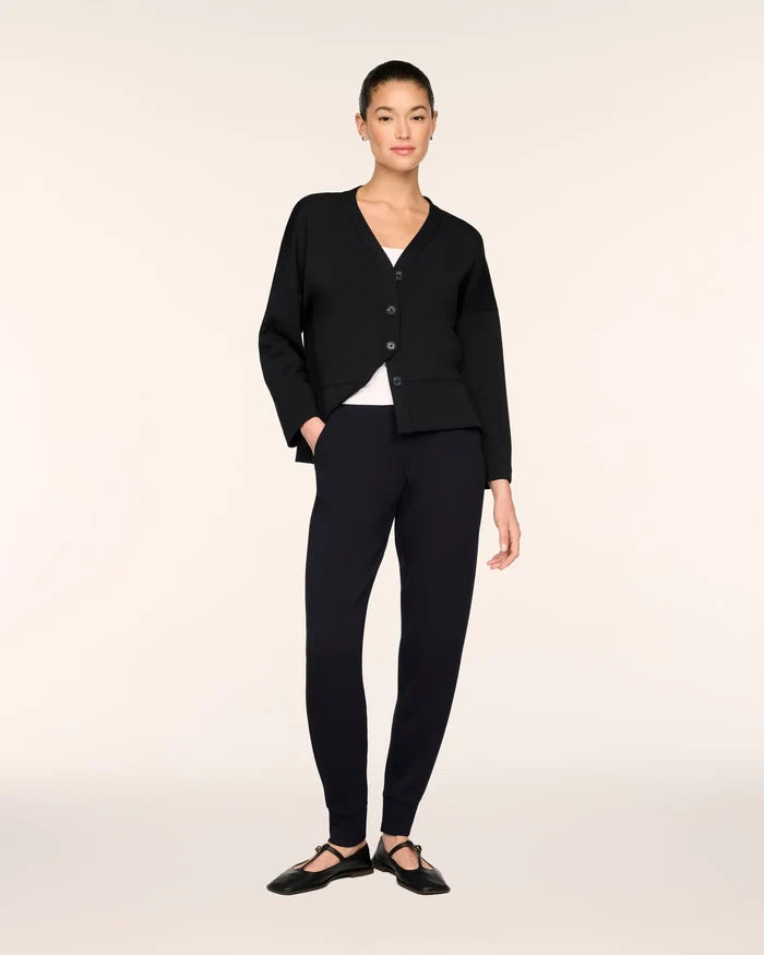 A model stands against a plain light beige studio background wearing a black cardigan with buttons over a white top, black fitted pants, and black ballet flats.
