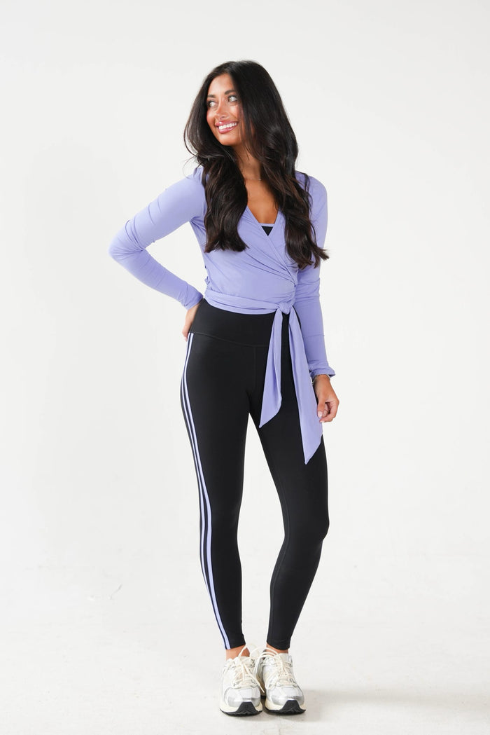 A woman modeling a light purple long-sleeve wrap top and black athletic leggings with white side stripes, paired with white sneakers, against a plain white studio background.