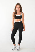 A woman modeling a matching black activewear set consisting of a sports bra with a white band and high-waisted leggings, paired with white sneakers, against a plain white studio background.