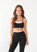 A woman modeling a matching black activewear set consisting of a sports bra with a white band and high-waisted leggings, paired with white sneakers, against a plain white studio background.