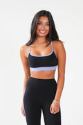 A woman modeling a black athletic sports bra and matching high-waisted leggings with light purple/lavender trim, photographed against a clean white studio background.