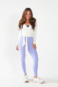 A woman modeling high-waisted light purple leggings with a white wrap-style top and white sneakers, standing against a plain white studio background.