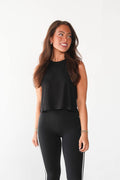A woman modeling a black sleeveless crop top and black leggings with white side stripes, paired with white sneakers, against a plain white studio background.