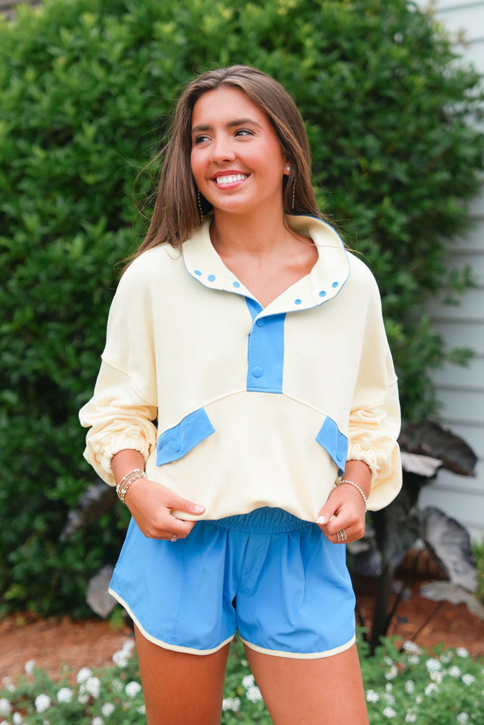 Woman wearing a yellow sweater with blue accents and blue shorts standing outdoors.