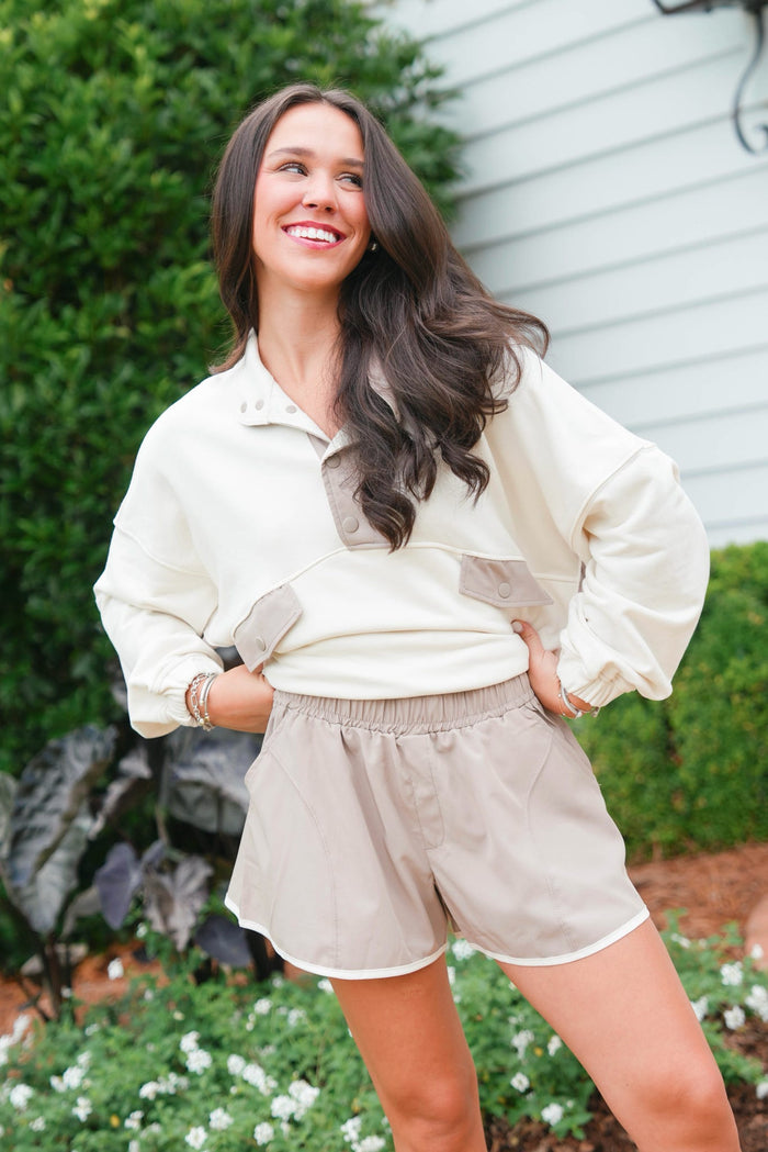 Woman wearing a cream jacket and beige shorts standing outdoors with greenery in the background