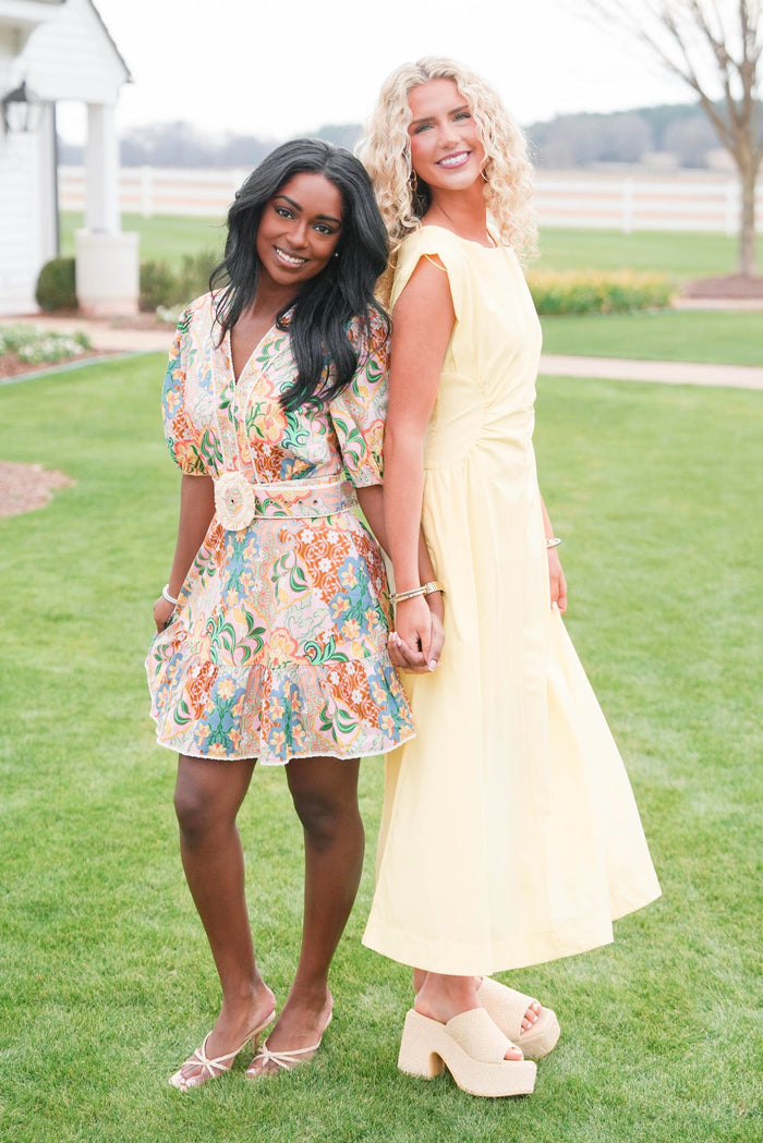 Two women standing on grass, one in a floral dress and the other in a yellow dress.