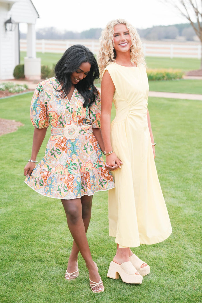 Two women standing on grass, one in a floral dress and the other in a yellow dress.
