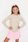 A young girl smiling and posing with her hands on her hips, wearing a long-sleeve floral print top with a pink bow headband and a pink skirt, set against a plain white background.