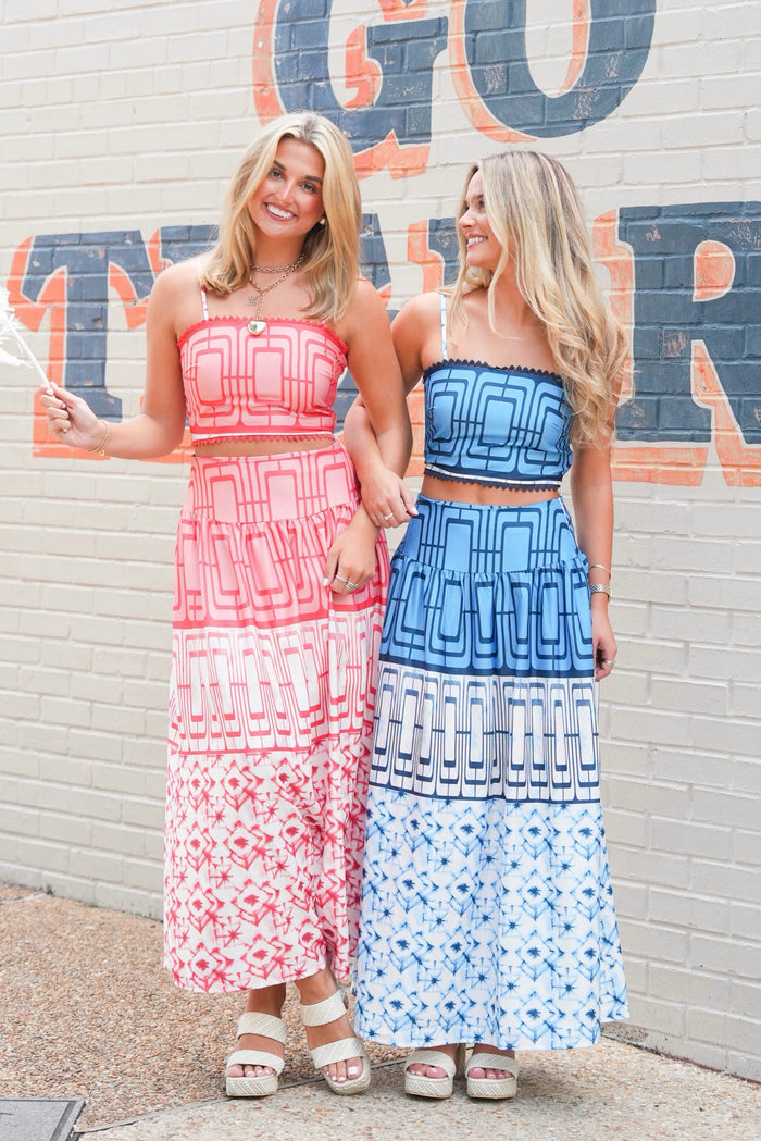 Two women standing side-by-side in matching geometric print maxi dresses, one in pink and one in blue, posing in front of a brick wall with painted lettering.