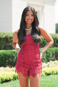 A woman wearing a bright red lace sleeveless romper stands outdoors in front of a white building and green hedges.