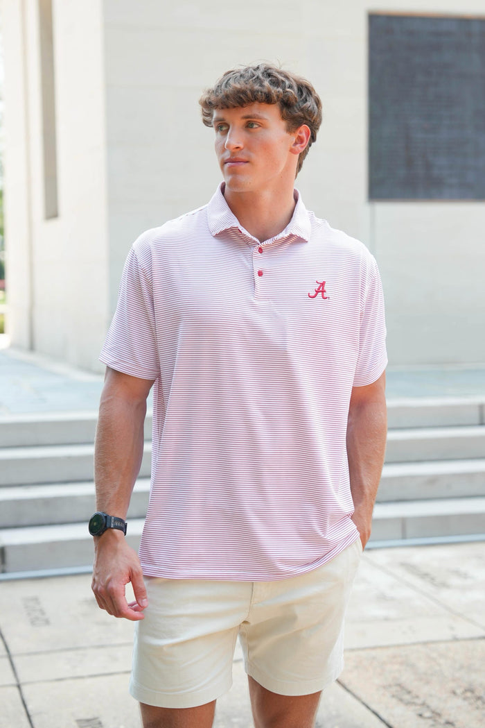 A man wearing a light pink and white horizontally striped polo shirt with a red Alabama 'A' logo on the chest, paired with beige shorts, standing outdoors in front of a building.