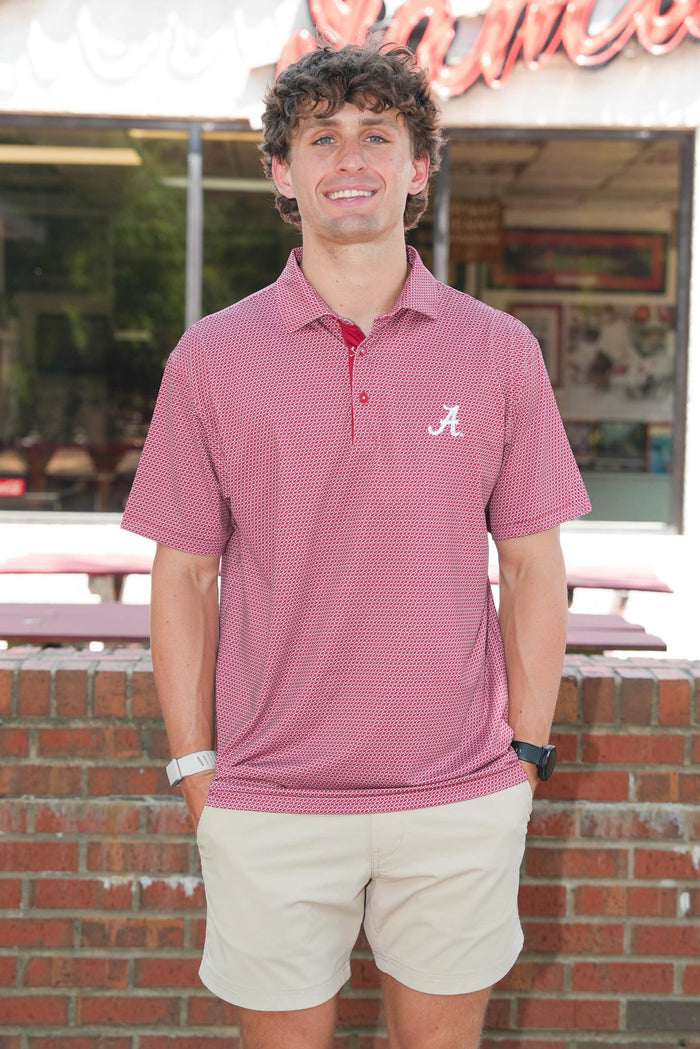 A man wearing a short-sleeve pink polo shirt with a white Alabama 'A' logo on the chest, paired with beige shorts, standing outdoors in front of a brick wall and storefront.