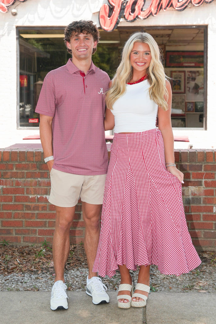 A man and a woman standing outdoors in front of a storefront. The man wears a dusty rose polo shirt, beige shorts, and white sneakers.