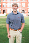 A smiling young man standing outdoors on a green lawn in front of a large red brick building, wearing a short-sleeve blue patterned polo shirt, khaki pants, and a brown belt.