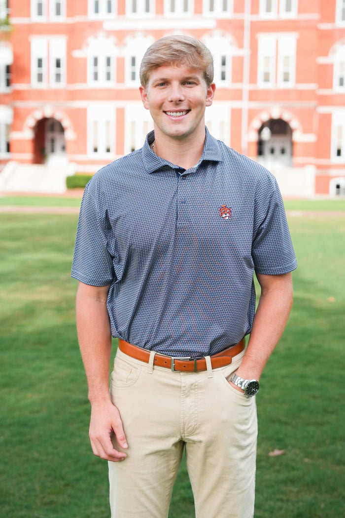 A smiling young man standing outdoors on a green lawn in front of a large red brick building, wearing a short-sleeve blue patterned polo shirt, khaki pants, and a brown belt.