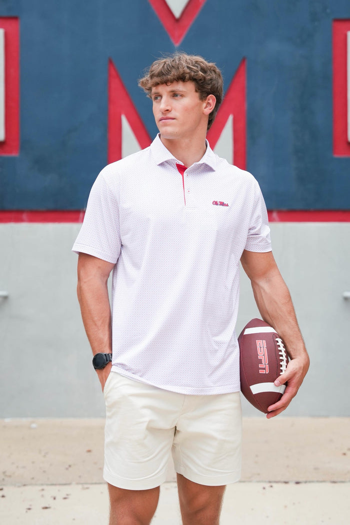 A man wearing a white short-sleeve polo shirt with a red collar and zipper, paired with cream shorts, holding a football against a blue and red wall background.