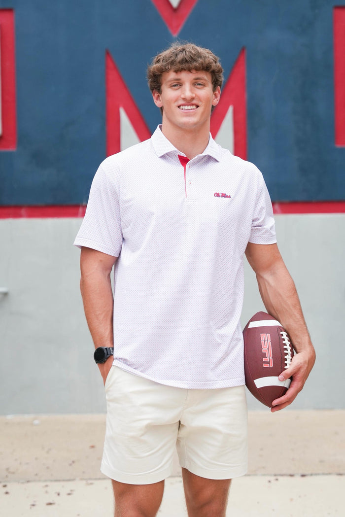A smiling young man stands outdoors holding a football, wearing a white short-sleeve polo shirt with a red collar and a small red Wilson logo on the chest, paired with cream shorts.