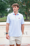 A young man wearing a light blue and white patterned short-sleeve polo shirt with a red zipper and a small red logo on the chest, paired with beige shorts, standing outdoors in front of a stone wall with trees in the background.