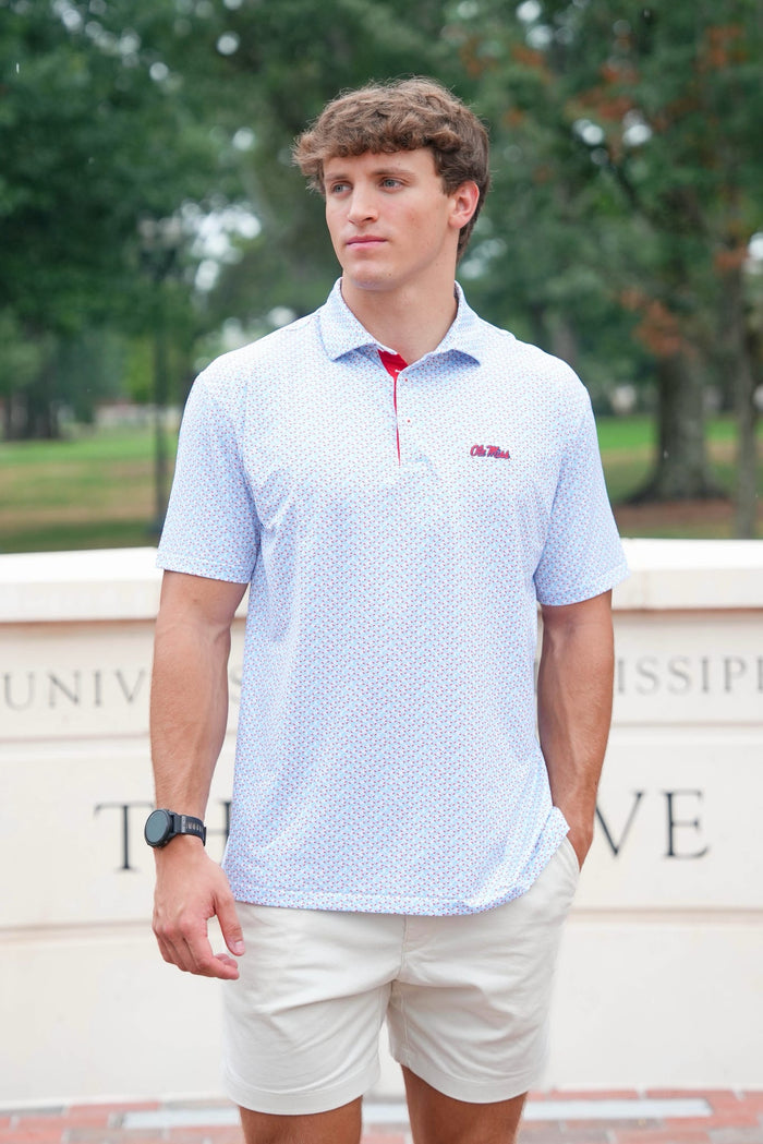 A young man wearing a light blue and white patterned short-sleeve polo shirt with a red zipper and a small red logo on the chest, paired with beige shorts, standing outdoors in front of a stone wall with trees in the background.