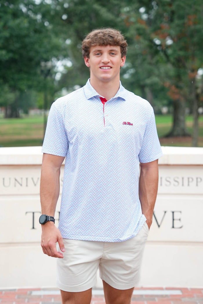 A young man smiling outdoors wearing a light blue and white patterned polo shirt with a red zipper and a small red logo on the chest, paired with beige shorts.
