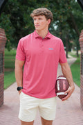 A man wearing a coral pink checkered polo shirt and cream shorts stands on a brick pathway holding a football. The polo shirt features a small embroidered logo on the left chest.