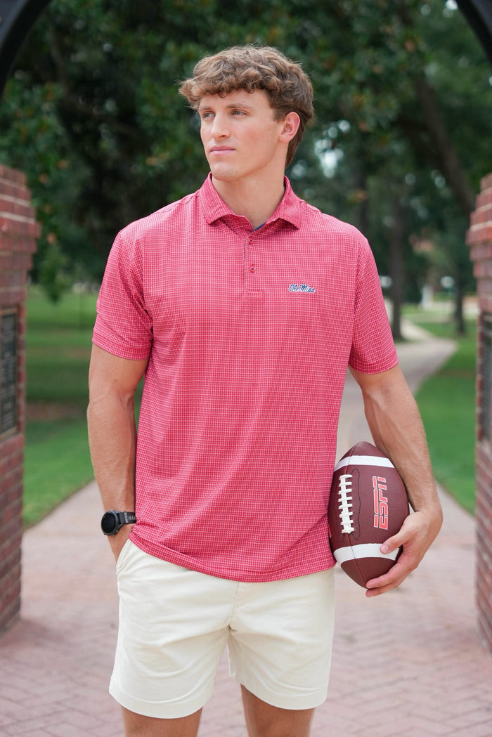 A man wearing a coral pink checkered polo shirt and cream shorts stands on a brick pathway holding a football. The polo shirt features a small embroidered logo on the left chest.