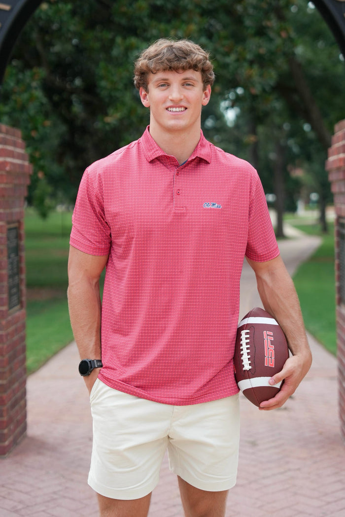 A man wearing a coral pink checkered polo shirt and cream shorts stands on a brick pathway holding a football. The polo shirt features a small embroidered logo on the left chest.