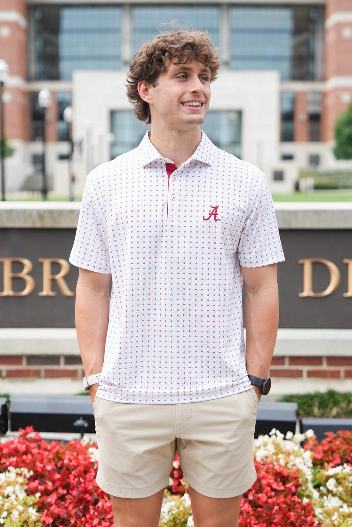 A man wearing a white short-sleeve polo shirt with a red micro-dot pattern, a red inner collar, and a red embroidered 'A' logo on the chest, paired with khaki shorts, standing outdoors in front of a brick building and flower beds.