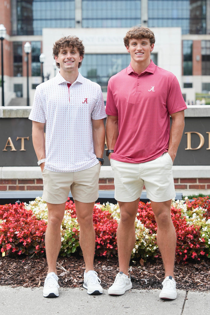 Two young men standing outdoors in front of a brick building and flowerbeds, wearing Alabama Crimson Tide themed polo shirts and khaki shorts. One wears a white patterned polo with a red 'A' logo, and the other wears a coral pink polo with a white 'A' logo.