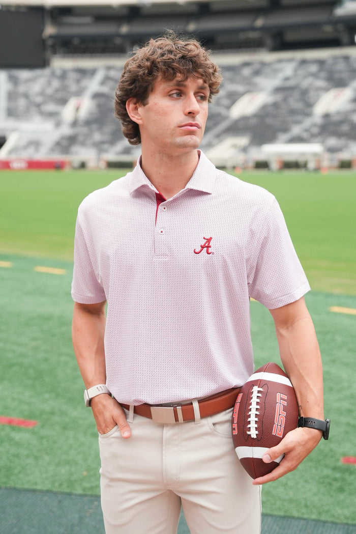 A man standing on a football field wearing a light pink polo shirt with a red Alabama 'A' logo on the chest, khaki pants, and holding a brown football.