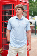 A man wearing a light blue and white striped polo shirt with beige pants, standing in front of a classic red telephone booth.