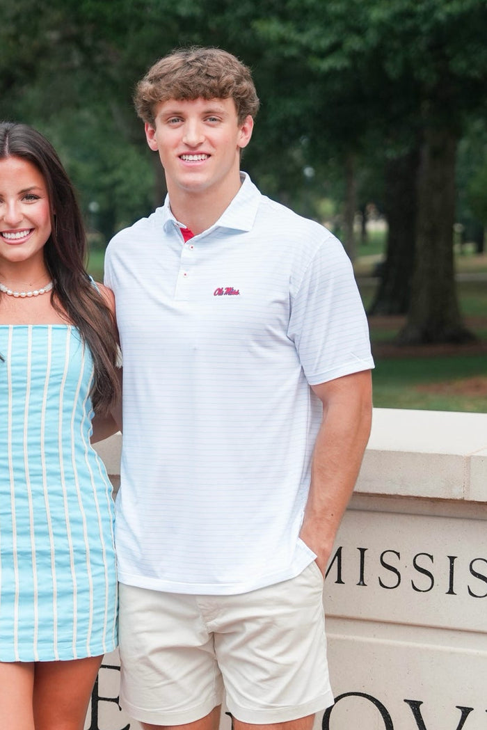 A young woman in a light blue and white striped mini dress and a man in a white polo shirt and beige shorts standing together outdoors in front of a brick and stone wall with green trees in the background.