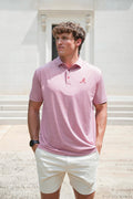 A man wearing a light pink short-sleeve polo shirt with a small red 'A' logo on the chest, paired with off-white shorts, standing outdoors in front of a light-colored building.