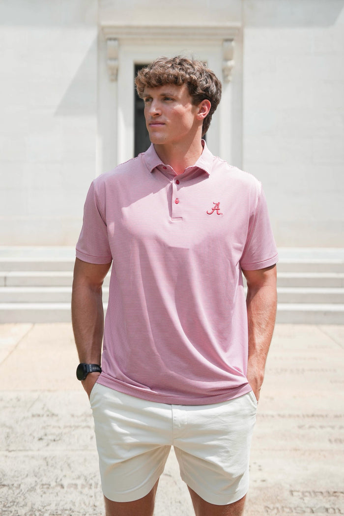 A man wearing a light pink short-sleeve polo shirt with a small red 'A' logo on the chest, paired with off-white shorts, standing outdoors in front of a light-colored building.