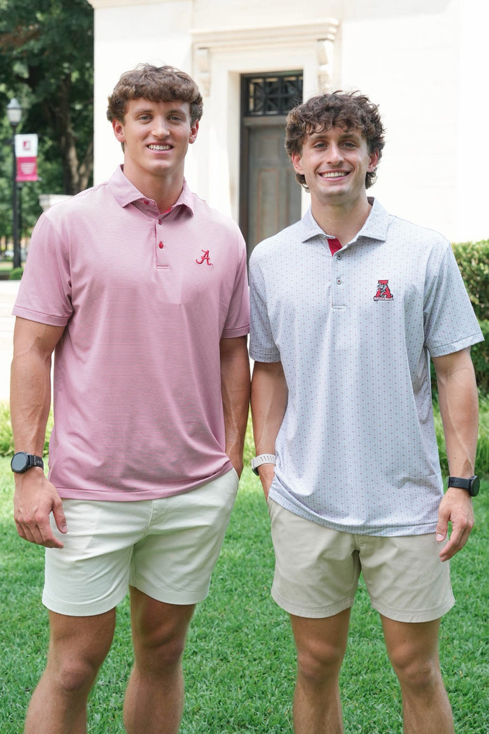 Two men standing outdoors on a grassy lawn in front of a building, each wearing a short-sleeve polo shirt with an Alabama logo on the chest. One shirt is light pink and the other is light gray, paired with khaki shorts.
