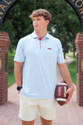 A young man stands outdoors holding a football, wearing a light blue polo shirt with a red collar and a small red Ole Miss logo on the chest, paired with cream shorts.