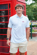 A man wearing a light blue polo shirt and beige shorts stands in front of a red telephone booth outdoors.