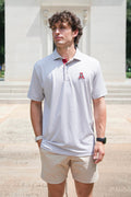 A man wearing a light gray and white patterned polo shirt with a red and white 'A' logo on the chest, paired with khaki shorts, standing outdoors in front of a stone building.