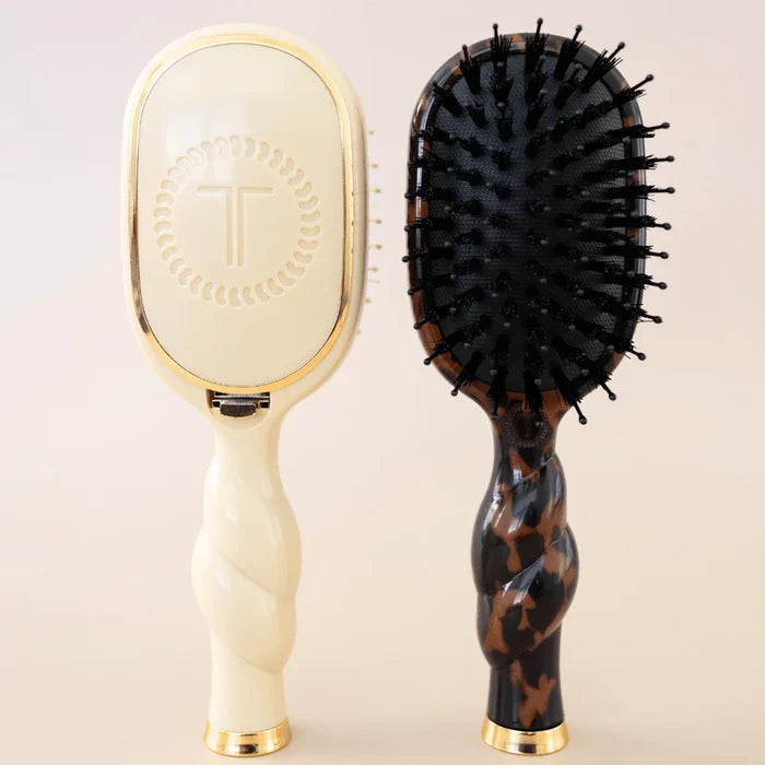 Two hairbrushes, one with a cream handle and gold accents, the other with a tortoiseshell pattern, on a light beige background.