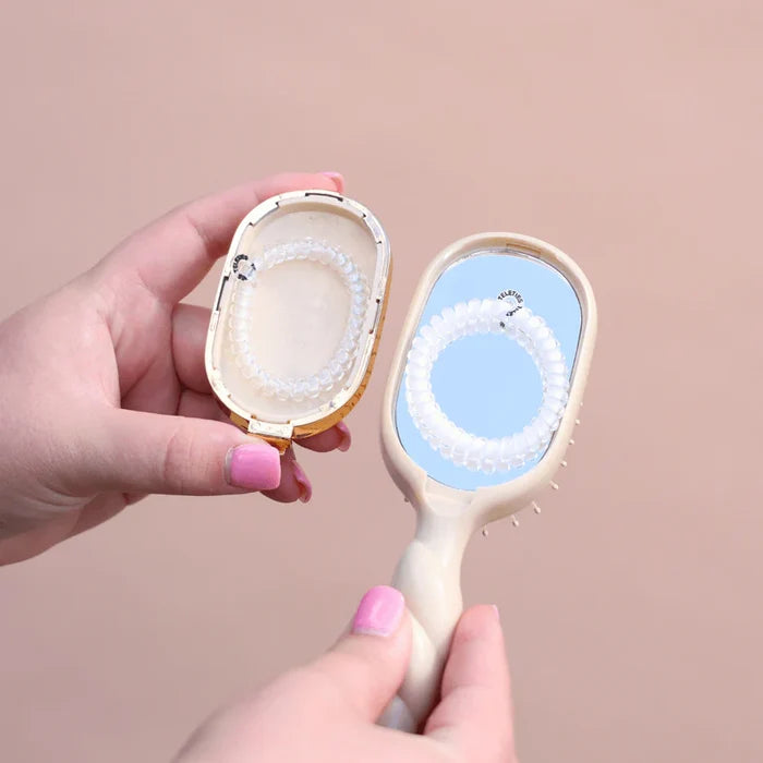 Hand holding a hair brush with compact mirror with the back of the brush with a small compartment holding 2 white 'Teletie' bands on a beige background