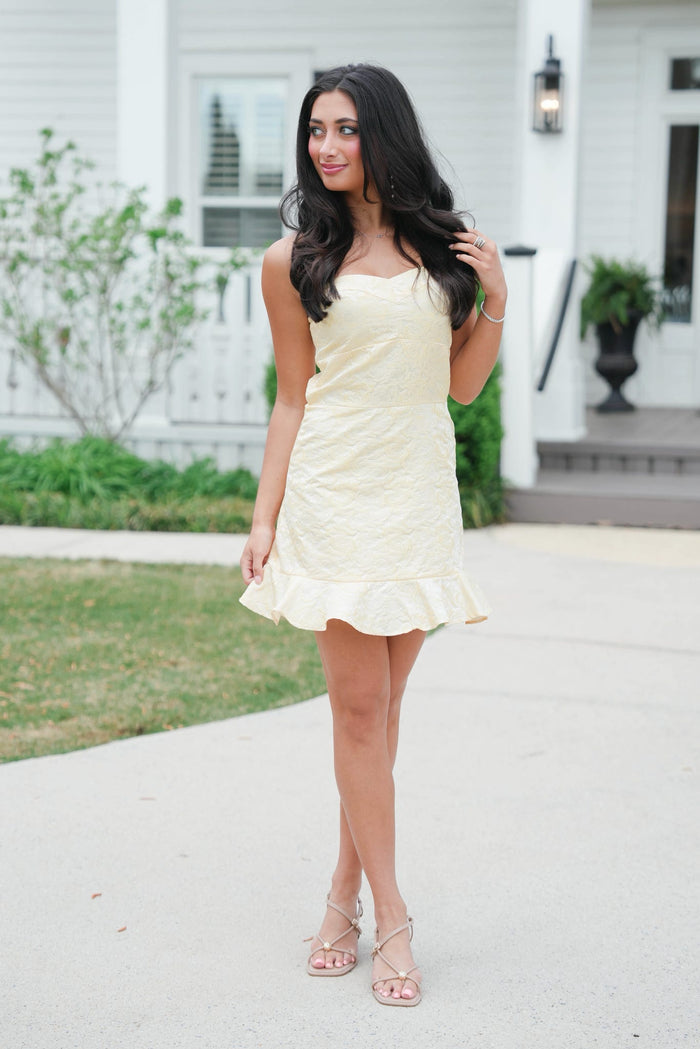 Woman in a light yellow dress standing on a sidewalk with a house in the background