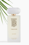 Thomas Blonde Blonde Eau de Parfum bottle with a frosted glass body, gold lettering, and a textured white cap, styled with green palm leaves on a white background.